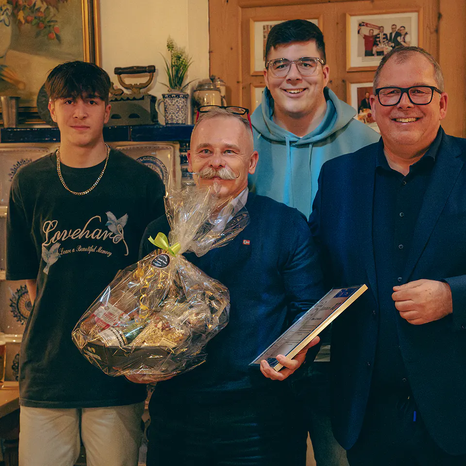 Vier Personen stehen nebeneinander und lächeln in die Kamera. In der Mitte hält ein Mann mit grauen Haaren und Schnurrbart einen großen Präsentkorb in Folie sowie eine Urkunde in der Hand. Links von ihm steht ein junger Mann, rechts daneben zwei weitere Kollegen. Alle wirken fröhlich und festlich gekleid
