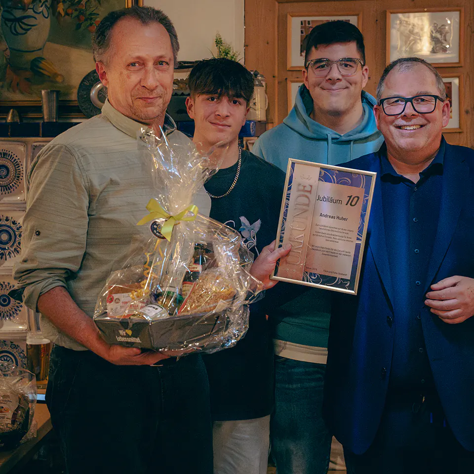 Andreas Huber – ein Mann im Hemd steht mit einem großen Präsentkorb in den Armen. Er eine Urkunde mit der Aufschrift „Jubiläum 10“. Zwei jüngere Begleiter und ein weiterer Mann im Anzug stehen lachend daneben.
