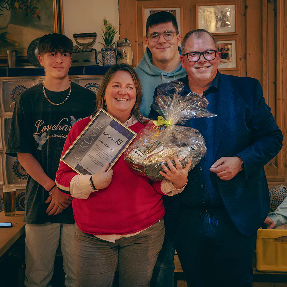 Claudia Sahlmann - eine Frau im roten Pullover hält stolz einen Präsentkorb in Folie sowie eine Urkunde mit „Jubiläum 15“. Sie strahlt und wird von drei Männern flankiert, darunter einer im Anzug.