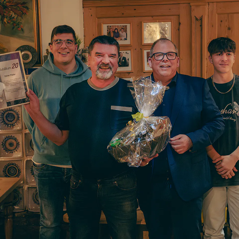 Ein Mann mit Bart im dunkelblauen T-Shirt lächelt in die Kamera. In der einen Hand hält er einen Präsentkorb, in der anderen eine Urkunde mit der Aufschrift „Jubiläum 20“. Neben ihm steht ein Mann im Anzug, hinter beiden zwei jüngere Begleiter.