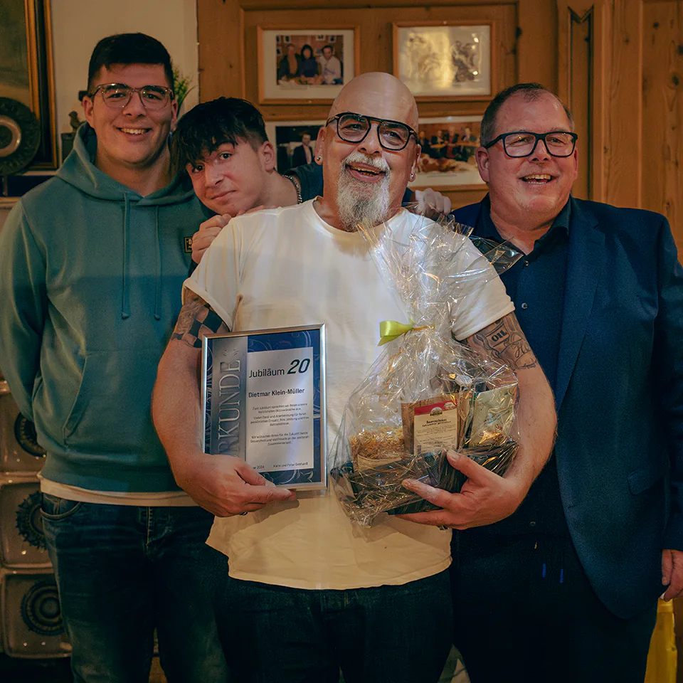 Ein Mann mit Glatze, Brille und weißem T-Shirt lacht herzlich. Er hält eine Urkunde für 20 Jahre Zugehörigkeit sowie einen großen Präsentkorb. Neben ihm steht ein Kollege im Anzug, hinter den beiden zwei jüngere Begleiter.
