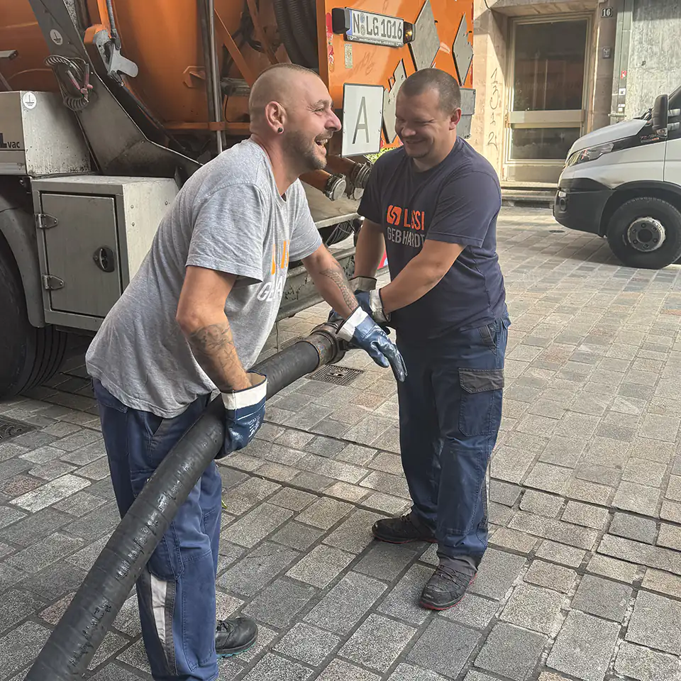 Zwei Mitarbeiter von LISSI GEBHARDT lachen und arbeiten gemeinsam mit einem Saugschlauch an einem Entsorgungsfahrzeug. Symbol für Teamgeist, Freude an der Arbeit und Zusammenarbeit im Alltag.
