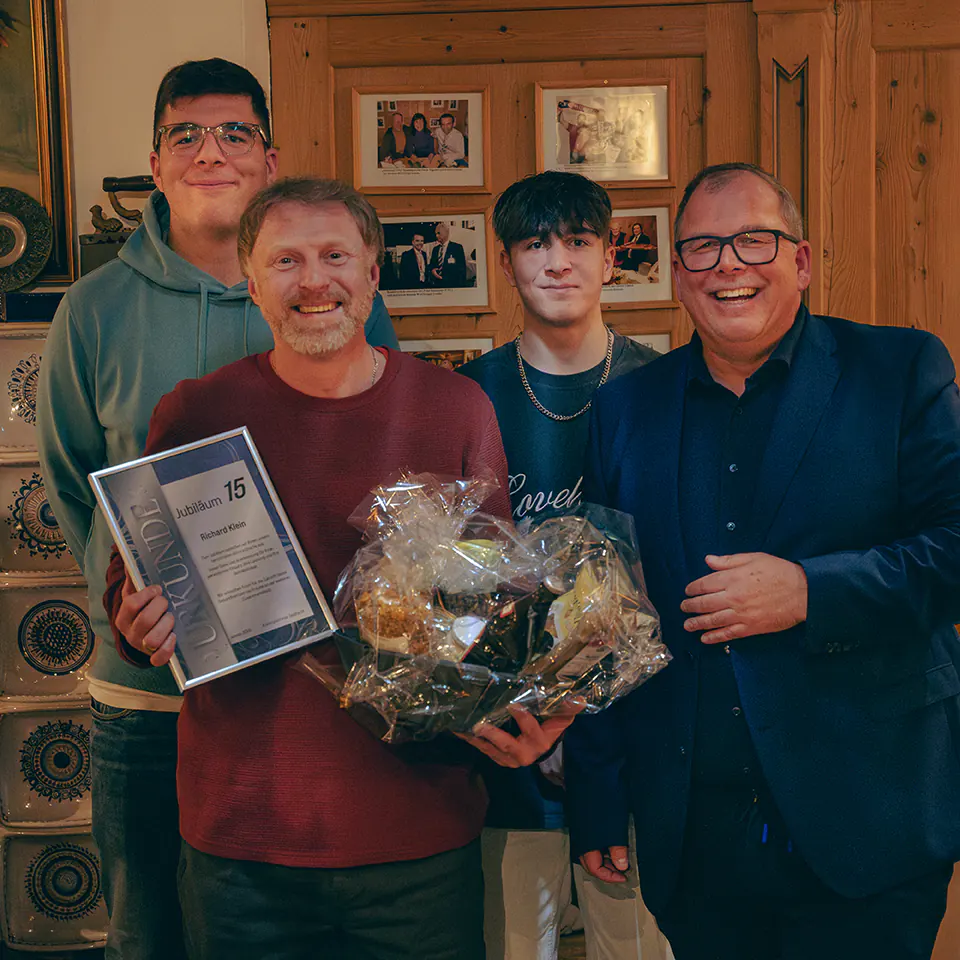 Ein Mann im roten Pullover strahlt und hält eine Urkunde „Jubiläum 15“ sowie einen großen Präsentkorb. Rechts daneben ein Kollege im blauen Anzug, hinter beiden stehen zwei jüngere Begleiter.